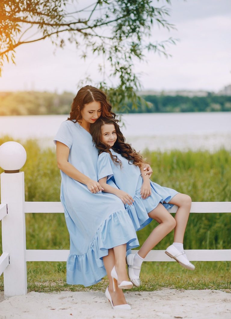 Achtsame Familienbegleitung in Dörpen Eine Frau und ein Mädchen in blauen Kleidern sitzen am Ufer eines Gewässers, auf einem Zaun. Die Frau hält das Mädchen liebevoll im Arm.
