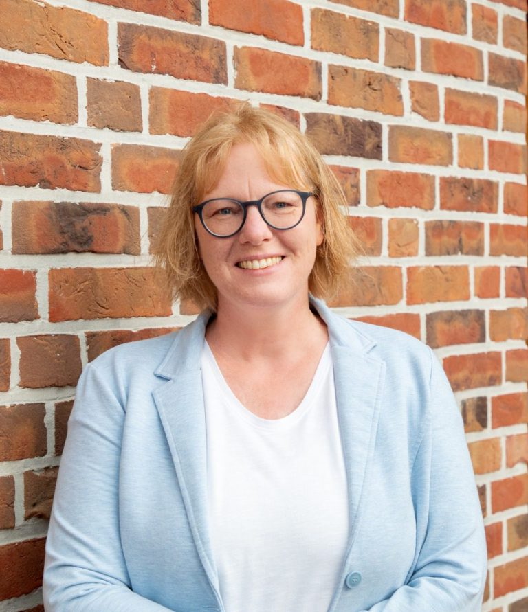 Achtsame Familienbegleitung in Dörpen Frau mit blonden Haaren und Brille, lächelt vor einer Backsteinwand.